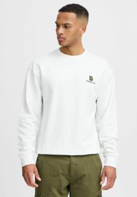 Balts džemperis ar garajām piedurknēm un apaļu izgriezumu. Iezīmēts ar mazu zaļu logotipu uz krūtīm. Mīksta tekstūra un brīvs griezums.