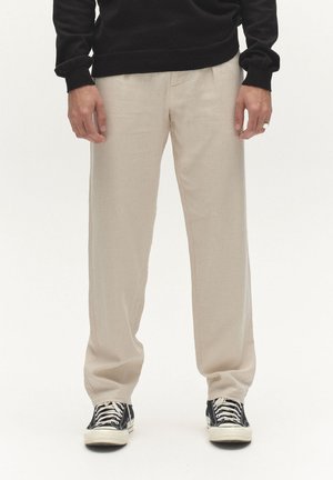 Mand iført beige bukser med lige ben, sort sweater og sort-hvide sneakers, stående mod en ensfarvet baggrund.