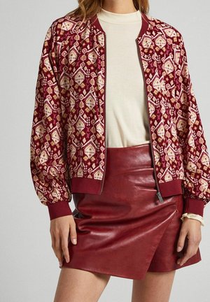 Femme portant une veste zippée bordeaux à motifs, un haut crème et une jupe en cuir bordeaux asymétrique, debout les mains relâchées le long du corps.