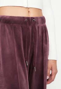 Nahaufnahme einer Person, die plumfarbene Samt-Trainingshosen und einen zusammengeschneiderten weißen Strickpullover trägt. Der Bauch ist sichtbar und die rechte Hand ist entspannt an der Seite.