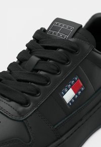 Close-up de um sapatilha preta com atacadores grossos e um emblema da Tommy Jeans em vermelho, branco e azul na lateral.