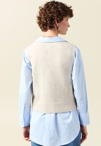 Beige Strickweste mit geripptem Saum über einem hellblauen Hemd mit Knopfverschluss, das lange Ärmel und einen klassischen Kragen hat. Strukturierter Stoff.