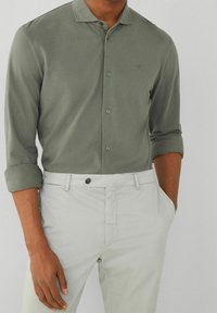 Chemise à manches longues vert olive foncé avec un col boutonné et un détail de logo subtil, associée à un pantalon beige clair. Texture lisse.