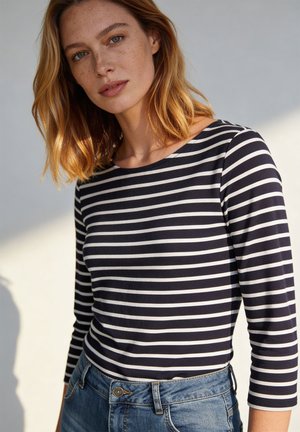 Jonge vrouw met rood haar en sproeten, gekleed in een zwart-wit gestreept shirt met lange mouwen en een blauwe spijkerbroek, staand tegen een neutrale achtergrond.