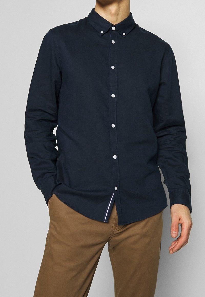 Uomo che indossa una camicia a maniche lunghe blu scuro abbottonata con colletto e pantaloni marrone chiaro, in piedi con una mano in tasca.