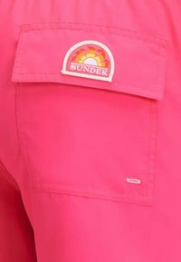 Tessuto rosa con una tasca a toppa caratterizzata da un colorato logo SUNDEK; texture liscia, design semplice, senza ulteriori fantasie o accessori.