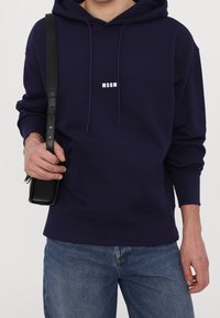 Sudadera azul marino de algodón, con un bolsillo frontal y texto de logo en blanco. Combinada con unos vaqueros azul claro y un bolso cruzado negro.