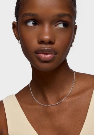 Primer plano de una persona con piel oscura y suave que lleva pequeños pendientes de aro plateados, un delicado collar de cadena plateada y una blusa sin mangas beige.