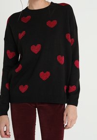 Musta neulepaita, jossa löysä istuvuus ja punaisilla kimaltavilla sydänkuvioilla koristeltu, resorit hihoissa ja helmassa. Yhdistetty viininpunaisiin housuihin.