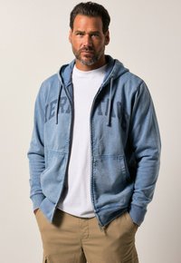 Zip-up hooded sweatshirt in light blue, featuring a front graphic print "HERITAGE," with two side pockets and a drawstring hood.