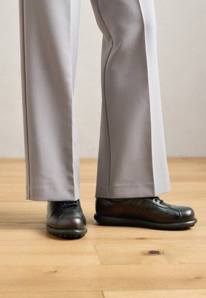 Des chaussures en cuir noir à lacets portées avec un pantalon large gris clair sur un sol en bois.