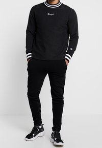 Felpa nera in cotone con polsini a righe bianche e nere e colletto a girocollo, abbinata a pantaloni jogger neri e sneakers nere. Logo Champion in evidenza.