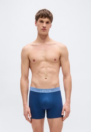 Νεαρός άνδρας με κοντά μαλλιά, φορώντας μπλε μποξεράκια Levi's, στέκεται με φόντο ένα απλό ανοιχτό γκρι φόντο, κοιτάζοντας προς τα εμπρός.