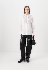 Chemise blanche à boutons avec un détail floral texturé sur la poitrine, associée à un pantalon en cuir noir ample et un petit sac à main noir.