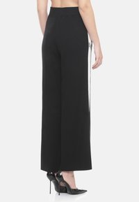 Pantaloni neri a gamba larga con una vita a coste, dotati di strisce bianche sui lati e un sottile dettaglio del logo lungo la cucitura esterna. Tessuto liscio.