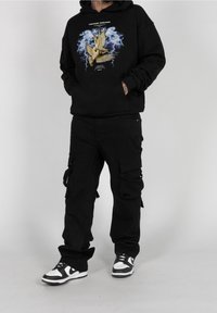 Black hoodie with graphic design of birds and lightning, paired with black cargo pants with pockets and straps, and black-and-white sneakers.