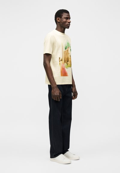 Άνδρας σε προφίλ φορώντας κρεμ κοντομάνικο με σχέδιο, μαύρο τζιν και λευκά αθλητικά παπούτσια, με απλό λευκό φόντο.