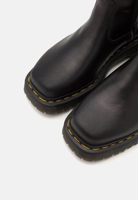 Black leather ankle boots with a square toe, yellow stitching along the sole, and a textured rubber outsole. Smooth surface and sturdy design.