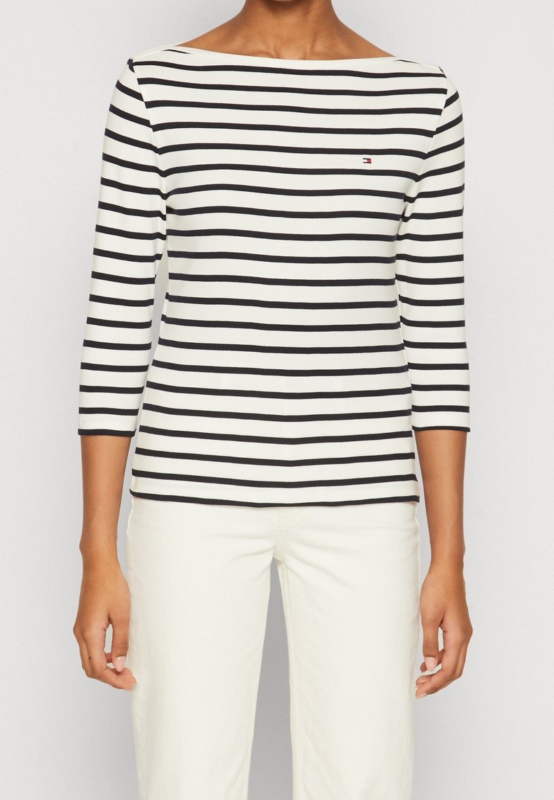 Bluzka z długim rękawem w paski w białym i czarnym kolorze, z dekoltem łódkowym i małym logo na piersi, zestawiona z białymi spodniami.