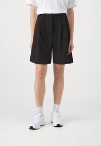 Bermuda shorts neri a pieghe realizzati in un tessuto morbido, caratterizzati da una vita alta e tasche laterali, abbinati a scarpe da ginnastica bianche e calze.