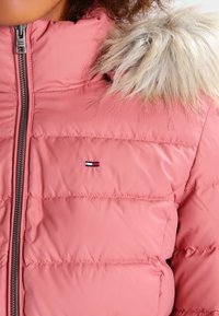 Rosa vadderad jacka med dragkedja, utrustad med huva fodrad med päls, sydda paneler och en liten logotyp på bröstet.