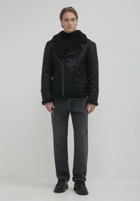 Giovane uomo in piedi dritto con giacca in shearling nera, dolcevita nero, jeans grigio scuro e scarpe di pelle nere su sfondo semplice.
