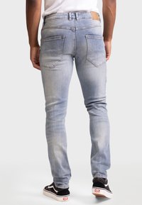 Vaqueros de mezclilla azul claro, corte ajustado, con cinco bolsillos, detalles sutiles de desgaste y una etiqueta de cuero en la cinturilla. Combinados con zapatillas negras.