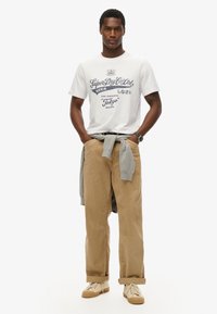 Camiseta de algodón blanca con texto azul, combinada con pantalones de pana beige y un suéter gris anudado a la cintura. Zapatillas beige completan el look.