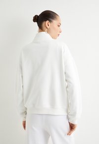 Sudadera blanca de gran tamaño con cuello alto, puños y cintura acanalados. Textura suave, costuras mínimas y diseño limpio.