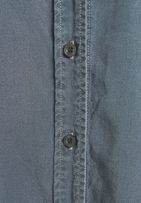 Jasnoniebieska koszula z tkaniny z ciemnymi guzikami i kontrastującym przeszyciem wzdłuż plisy, o gładkiej fakturze i lekko pomarszczonym wyglądzie.