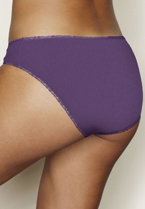 Frau trägt lila Spitzen-Slips, Nahaufnahme von Hüfte und oberem Oberschenkel vor einfarbigem hellem Hintergrund.