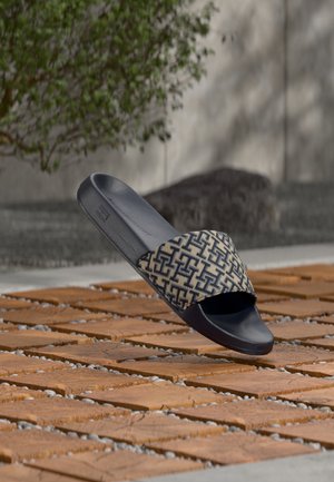 Sandalia de diapositiva negra con una tira de tela beige y negra con patrón, flotando sobre una superficie exterior de ladrillos y guijarros.