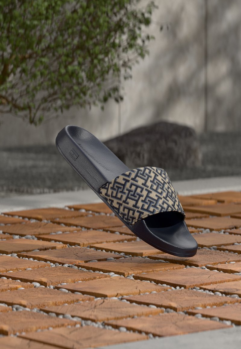 Mule noire avec bride en tissu beige et noir à motifs, flottant au-dessus d'une surface extérieure en briques et cailloux.