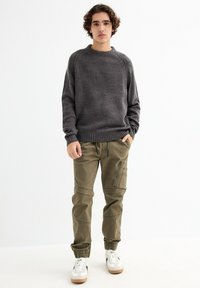 Suéter de punto gris oscuro con cuello redondo, combinado con pantalones cargo oliva que cuentan con cremalleras y puños. Zapatillas blancas completan el look.