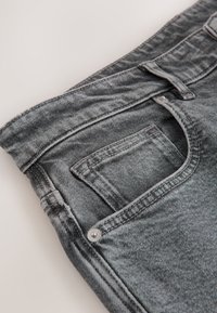 Tuvplāns ar izbalējušām melnām džinsa biksēm, kur redzams priekšējais kabata, mazā monētu kabata, jostas cilpa un metāla kniedes detaļas.