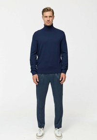 Sötétkék garbó pulóver texturált sötétszürke nadrággal. Az öltözet jól illeszkedő kivitelű, és fehér sportcipővel párosított.