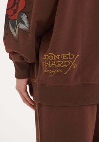 Hnědá mikina s velkým grafickým motivem červené růže a zlatým nápisem "Don Ed Hardy designs" na lemu. Podrobnosti s ribbovanými manžetami a pasem.