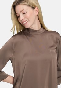 Blusa di seta marrone con collo alto e maniche 3/4, caratterizzata da una texture liscia e una finitura leggermente lucida, progettata per un fit rilassato.