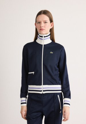 Veste de survêtement marine avec un col haut, accents rayés blancs et violets, fermeture éclair sur le devant et un petit logo vert. Tissu doux et extensible.
