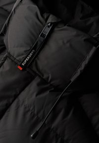 Sort dunjakke med en glat, mat finish, udstyret med lynlås og snøre. "SUPERDRY" logoetiket i rød og hvid detalje.