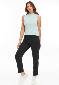 Helles blaues, ärmelloses Oberteil mit hohem Kragen, kombiniert mit hochgeschnittenen schwarzen Jeans und weißen Sneakers. Glatte Stricktextur.