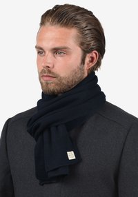 Sötétkék kötött sál, puha anyagból, texturált mintával és egy kis márkajelzéssel ellátott címkével, egy modell nyakán drapírozva.