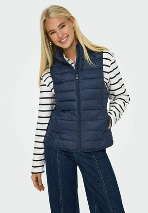 Gilet matelassé bleu marine avec un design quilté, fermeture éclair à l'avant, col montant, porté par-dessus une chemise rayée blanche et bleu marine, associé à un jean foncé.