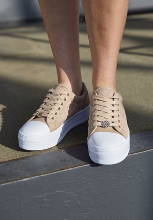 Beżowe sneakersy z koronkowymi detalami i białą grubą podeszwą, noszone na bosych stopach, stojące na krawędzi zewnętrznego chodnika w świetle słonecznym.