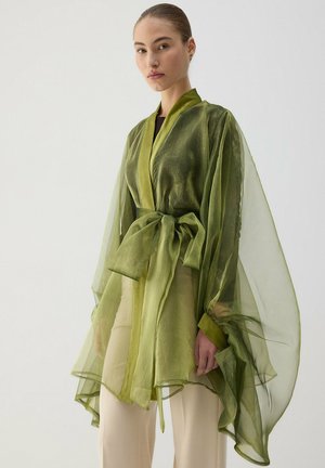 Jeune femme portant une veste portefeuille olive transparente avec un grand nœud de ceinture, associée à un pantalon large crème sur un fond clair uni.