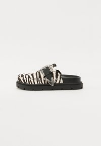 Black and white zebra-patterned slip-on shoe with a thick black sole, highlighted by a decorative silver buckle and studded accents.