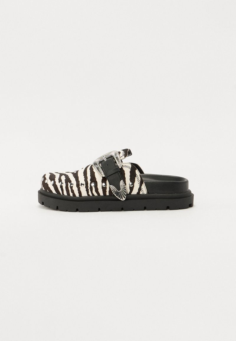 Black and white zebra-patterned slip-on shoe with a thick black sole, highlighted by a decorative silver buckle and studded accents.