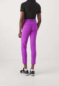 Person wearing fitted purple checkered pants, black short-sleeve shirt, white socks, and black sneakers standing against a plain light background.