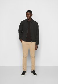 Zwarte bomberjack met rits, gecombineerd met een donker shirt, beige chinos en zwarte sneakers. Rechtermode ontwerp met een minimalistische stijl.