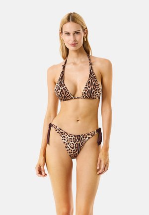 Donna con capelli biondi che indossa un bikini marrone stampa leopardata con laccetti ai lati e orecchini a cerchio su uno sfondo chiaro uniforme.